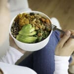 woman eating a bowl of healthy food including avocado and cauliflower and other vegetables.