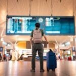 Traveler standing in an airport terminal with a backpack and suitcase, looking up at a large departures board.