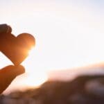 Hand holding a small heart-shaped object against the setting sun near a body of water.