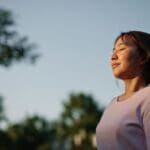 Woman standing outdoors with eyes closed, appearing calm and peaceful as sunlight gently illuminates her face.