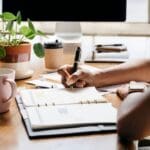 Person writing in a planner at a desk with coffee mugs, papers, and a potted plant, symbolizing organization and planning.