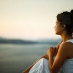 A woman in a white dress sits outdoors at sunset, gazing thoughtfully over the water, suggesting reflection, peace, or contemplation.