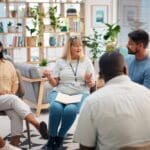 Group of people sitting in a circle during a therapy session, with a facilitator leading the discussion in a bright, supportive setting.