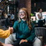 Smiling woman in glasses shaking hands with another person in a professional setting, suggesting a new job or successful meeting.