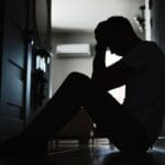 Silhouette of a person sitting on the floor with head in hands, appearing distressed or overwhelmed.