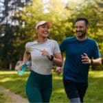 Man and woman jogging together in a sunny park, smiling and enjoying their run while holding a water bottle.