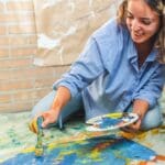 Smiling woman sitting on the floor painting a colorful canvas, enjoying a creative hobby in a relaxed setting.