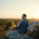 A man sits on a rocky hillside at sunset, peacefully reflecting while overlooking a vast landscape—symbolizing hope and grounding in recovery.