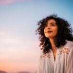 A woman with curly hair looks upward with a peaceful expression during sunset. Soft pastel colors fill the sky behind her, creating a calm, reflective mood.