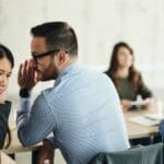 Two coworkers sit close together as one whispers to the other, while colleagues talk in the background, suggesting workplace gossip.