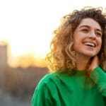 A woman smiling brightly outdoors at sunset, appearing joyful and relaxed.