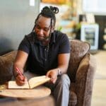 A man sits in a comfortable chair writing in a notebook, journaling quietly in a relaxed indoor setting.