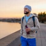 A man wearing a hoodie, beanie, and backpack walks along a waterfront path at sunset, smiling and holding a water bottle.