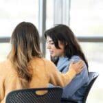Two people seated indoors, one comforting the other with a supportive hand on their back. Photo depicts good mental health practices in recovery.