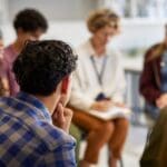 The Aviary - Group Therapy Provides a Great Group of Benefits Part One: Collective Wisdom and Experience. A group of individuals sit in a circle. A woman out of focus writes on a clipboard.